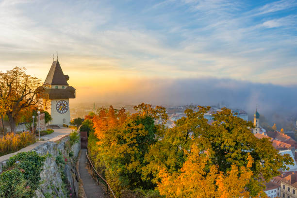 Clock tower atop a hill overlooking a city with autumn trees in orange and gold at sunrise/sunset.